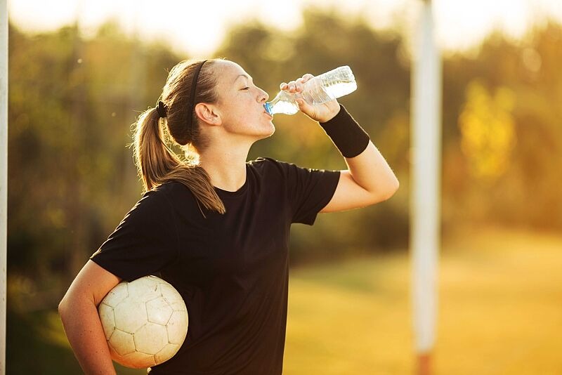 Eine Frau trinkt aus einer Wasserflasche und hält dabei einen Fußball.