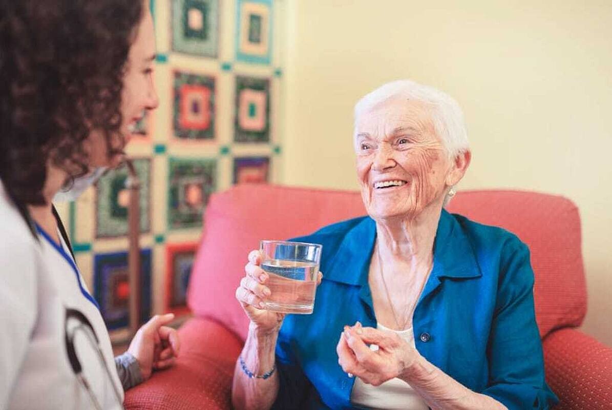 Eine ältere Frau hält ein Wasserglas in der Hand und lächelt eine Pflegekraft an.