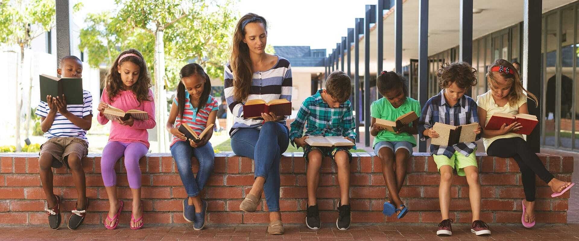Eine Lehrerin sitzt mit ihren Schülern und Schülerinnen im Innenhof der Schule auf einem Mäuerchen, jeder liest ein Buch
