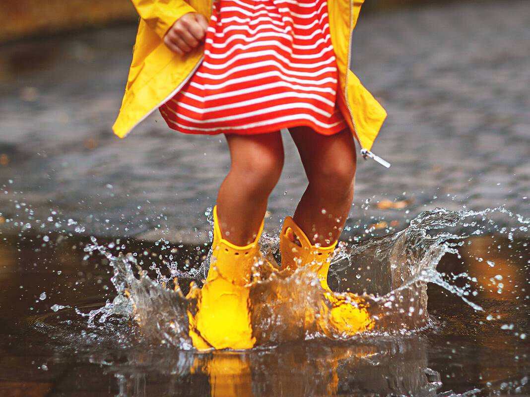 Foto eines Kindes in rot-weiß gestreiftem Kleid, gelber Regenjacke und gelben Regenschuhen, das in Pfütze springt